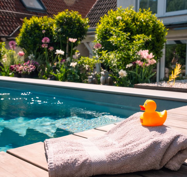 Sommerlicher Poolgarten mit Quietscheente Gartenpool mit Holzdeck, blühenden Pflanzen und einem Handtuch mit Quietscheente im Vordergrund, dahinter ein Wohnhaus mit großen Fenstern.